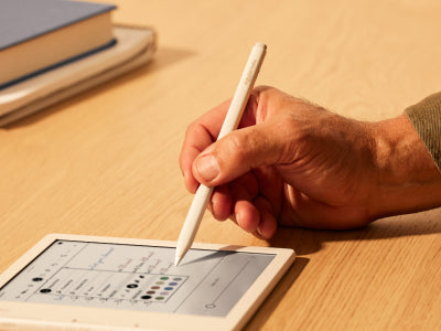 O mână este arătată ținând un Kobo Stylus 2 deasupra unui eReader Kobo Libra Colour pe o suprafață plană. Vârful stylus-ului este pe cale să selecteze una dintre culorile de stilou afișate într-un meniu de opțiuni pe ecranul eReader-ului.