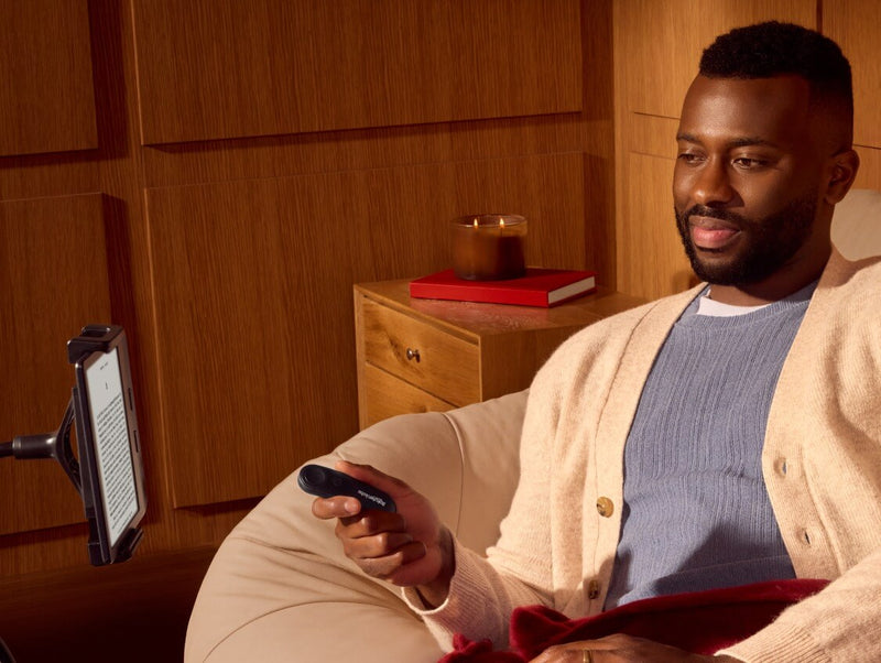 En man sitter bekvämt och håller Kobo Remote i höger hand medan han tittar på en Kobo e‑läsare på ett flexibelt stativ.