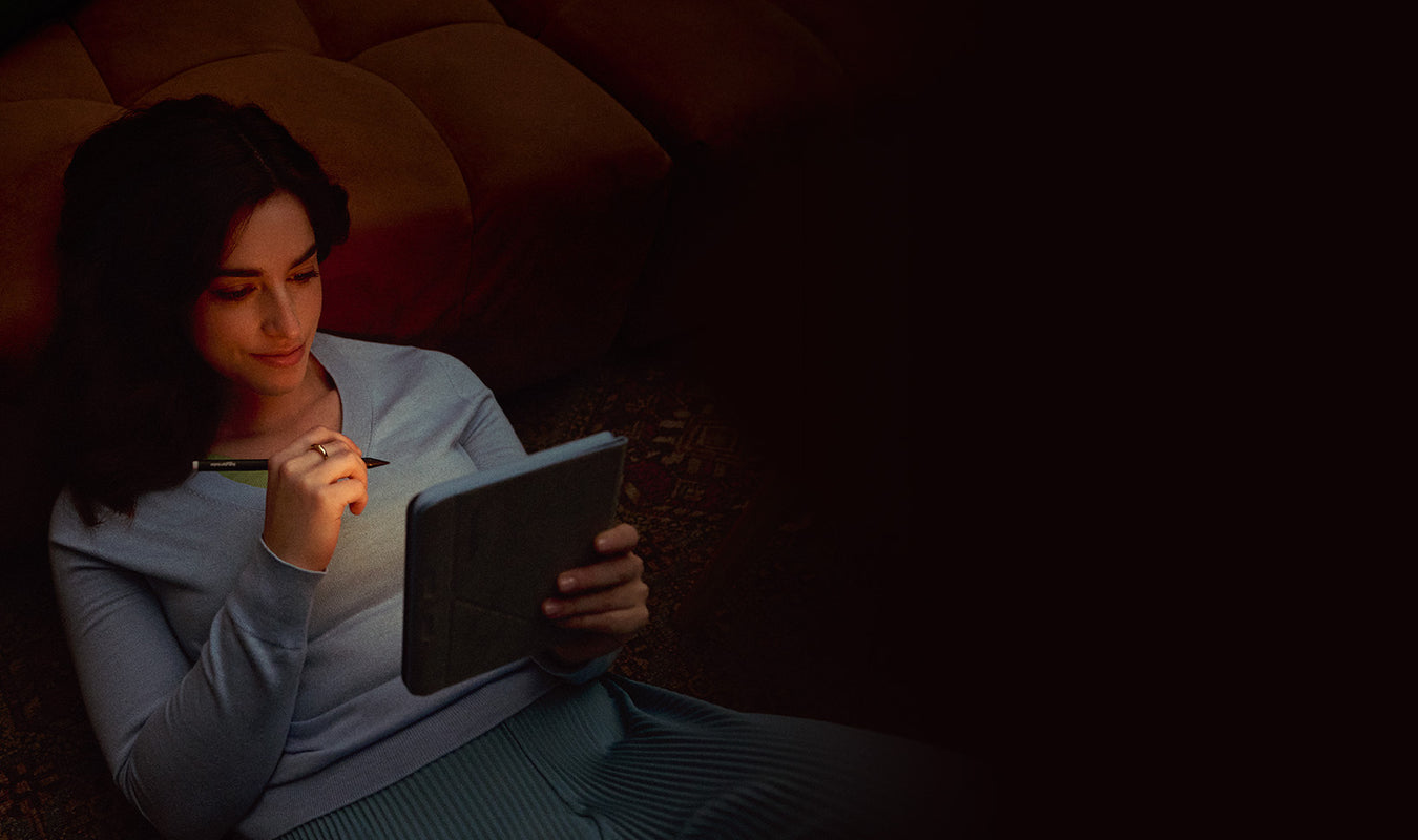A woman reclining on a couch, lit by the soft light of a Kobo Libra Colour eReader she is holding, along with a Kobo Stylus 2.